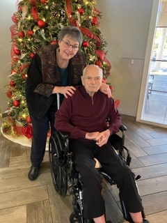 Photo of Diane R and her husband Larry in front of their Christmas tree, Diane smiling and leaning over Larry's wheelchair