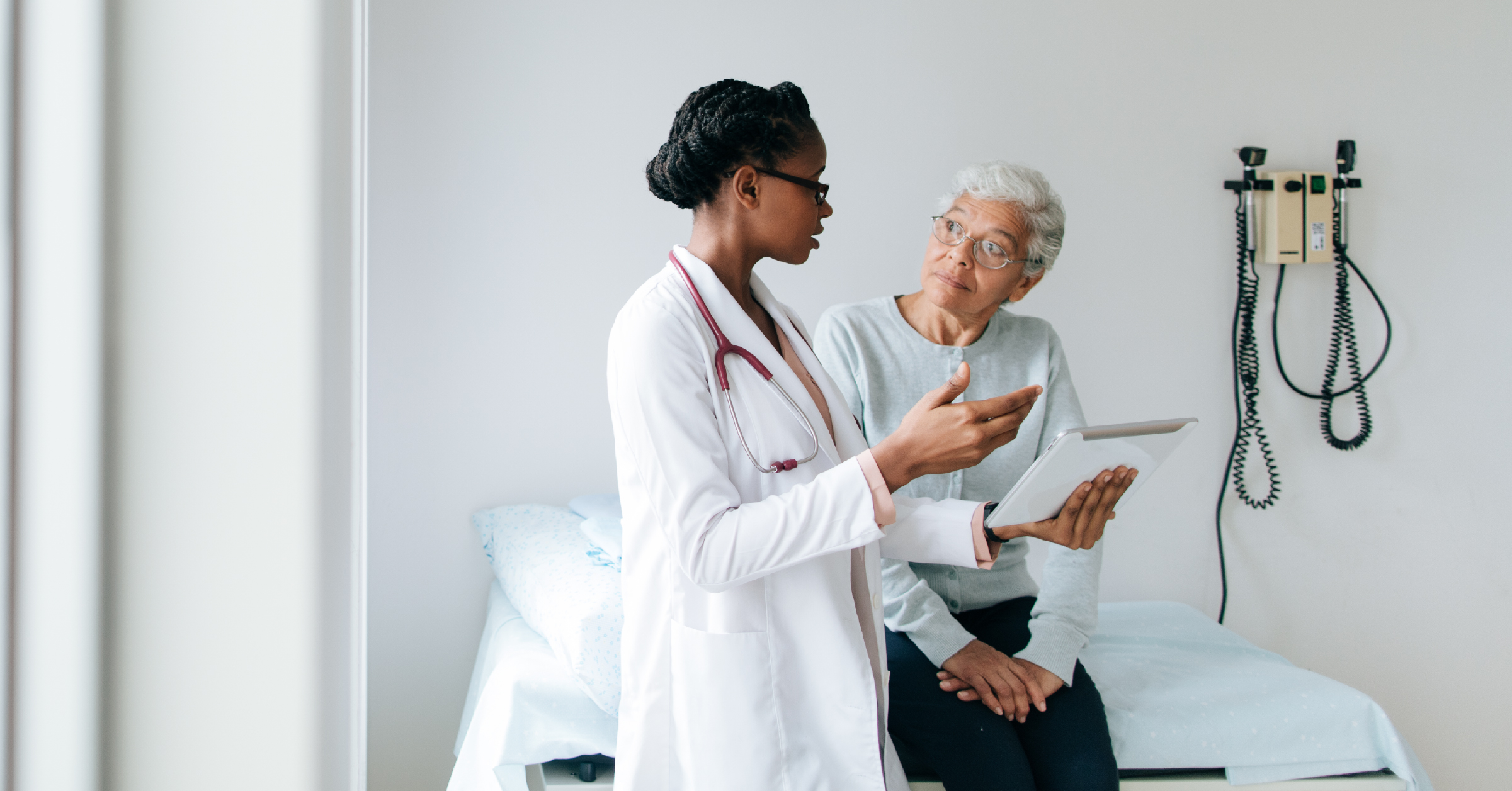 A photograph of an African American female doctor talking with a female patient of color, reading from a tablet device